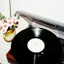 Als er das Lied vom Tod abspielte Ein Retro-Plattenspieler auf einem weißen Regal, aufgenommen vor einer weißen Wand. Neben dem Plattenspieler stehen eine kleine Vase mit rosa Blumen und ein gerahmtes Bild. Unter dem Regal sind Schallplatten und Bücher zu sehen.