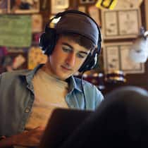 Das erste Mal mit 18 Ein Jugendlicher mit Kopfhörern und Mütze sitzt in seinem Schlafzimmer und arbeitet an seinem Laptop.