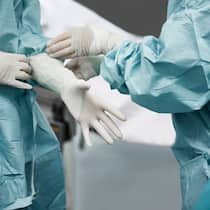 Profite fehlen, Patienten sterben Eine Ärztin hilft der anderen, Handschuhe anzuziehen. Beide tragen Schutzüberzüge. Man sieht nur die Arme und den Rumpf