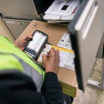 Paket verschwunden – was jetzt? Ein Pöstler steht mit einem Päckli vor einem Briefkasten, das nicht ins Ablagefach passt und schreibt deshalb eine Abholungseinladung an die Empfängerin.