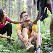 Tipps für eine sichere Wanderung Ein Wanderer mit Turnschuhen hat sich den Knöchel verstaucht, sitzt auf einem Waldboden und wird von vorbeilaufenden Wanderern angefragt, ob alles okay ist.