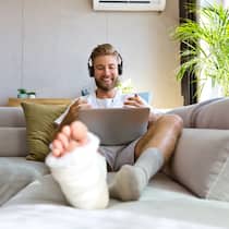 Mehr Rechte für Versicherte Ein junger Mann, dessen rechtes Bein mit einem Gips versehen ist, sitzt vergnügt auf dem Sofa während er einen Videoanruf mit seinem Laptop macht.