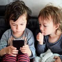 Kind kauft Spiele-Apps – müssen Eltern zahlen? Zwei kleine Jungs sitzen auf einem Sofa, der eine spielt mit dem Handy, der andere schaut ihm mit dem Finger nuckelnd dabei zu.