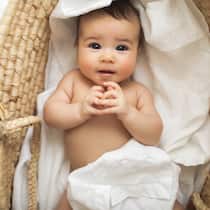 Auf Umwegen zum Baby Ein kleines Baby liegt umwickelt in weissen Laken in einem Korb und lächelt süss in die Kamera.