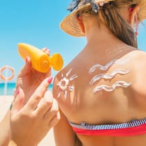 So vermeiden Sie hormonaktive Stoffe Ein kleines Mädchen am Strand steht mit dem Rücken zu ihrer Mutter, die ihr Sonnencreme in lustiger Wellen- und Sonnenform aufträgt.