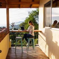 Das Büro sauber an den Strand zügeln Eine junge Frau sitzt auf der Terrasse ihrer Wohnung, die sich inmitten einer grünen Landschaft befindet. Was können Remote Workerinnen tun, damit ihr Arbeitsaufenthalt im Ausland klappt und das auch noch nachhaltig für Land und Leute?