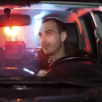 So schnell ist das Billett weg Ein junger Mann sitzt nachts am Steuer seines Auto, hinter ihm ist ein rot-blaues Licht eines Polizeifahrzeugs zu sehen.