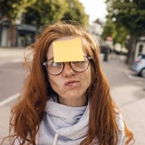 Wie viel Vergesslichkeit ist normal? Eine rothaarige Frau hat einen gelben Notizzettel auf ihre Stirn geklebt, um sich an etwas zu erinnern.