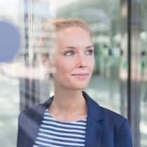 Selber entscheiden Eine Geschäftsfrau mit blauem Blazer schaut zufrieden und selbstbewusst durch ein verglastes Bürogebäude.