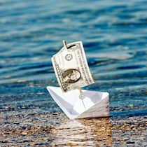 Sollen wir das Reisebüro betreiben? Ein Papierschiff mit einer Dollar-Note als Segel droht zu kentern und steht symbolisch für die USA-Rundreise, welche wegen der Pandemie storniert werden musste.