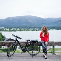 Worauf beim Kauf aus zweiter Hand achten Eine junge Frau sitzt bei ihrem Ausflug mit einem E-Bike auf einem Holzbacken am Strassenrand und trinkt während einer Pause von einer Wasserflasche.