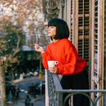 Wie mit schlechten Gewohnheiten aufhören? Eine junge Frau steht auf ihrem Balkon während einer Arbeitspause, die sie mit einer Tasse Kaffee und einer Zigarette verbringt.