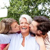 Muss ich jetzt mein Testament ändern? Zwei erwachsene Kinder küssen ihre lachende Mutter auf die Wange, während sie ein Selfie von ihnen zusammen im Garten macht.