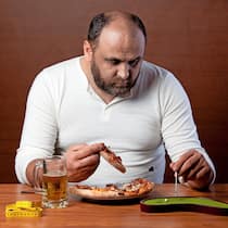 So entlasten Sie Ihre Leber Ein übergewichtiger Mann isst eine Pizza und spielt Tischgolf, um sich nicht bewegen zu müssen.
