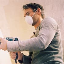 Krankmacher im Wohnzimmer Ein Mann mit Mundschutz schleift eine Wand ab.