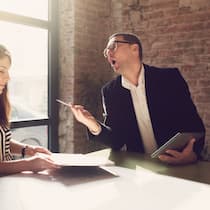Nervige Mitarbeiter entlassen? Zwei Angestellte sitzen in einem Besprechungszimmer. Der Angestellte rechts macht unmögliche Bemerkungen, welche die Mitarbeiterin zur linken nerven.