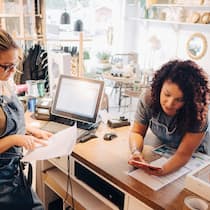 Wie Sie Ihre Liquidität langfristig sichern Zwei Geschäftsfrauen sind in ihrem Kleidergeschäft und beschäftigen sich mit Zahlen und Unterlagen.