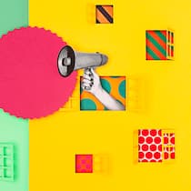 Muss ich laute Nachbarn ertragen? Eine grüne und eine gelbe Hauswand, aus deren Fenster eine Hand mit Megafon herausragt. Wie viel Lärm man unter Nachbarn ertragen muss.