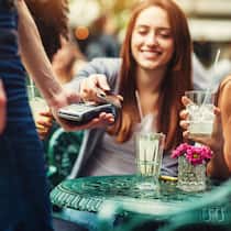 Endlich Geld – und jetzt? Zwei junge Frauen sitzen draussen in einem Café und bezahlen mit dem Handy für ihre Drinks.