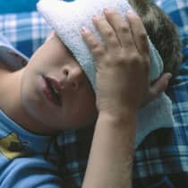 Jedes zehnte Kind hat Migräne Ein Junge liegt im Bett und hält sich einen Waschlappen vor die Augen.