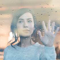 «Ich fühle mich so ausgelaugt» Eine junge Frau blickt in Gedanken versunken aus dem Fenster und hält ihre linke Hand gegen das Fensterglas, auf dem einzelne Regentropfen liegen.