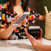 Wie steige ich auf Bargeldlos um? Eine junge Frau hält ihr Smartphone zum Bezahlen an ein Lesegerät.