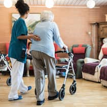 Worauf ist beim Umzug in ein anderes Pflegeheim zu achten? Pflegerin stützt alte Frau mit Rollator im Altersheim