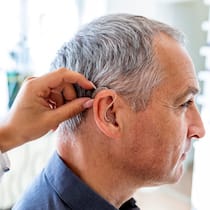 Ist mein Hörgerät bei Verlust versichert? Ein älterer Mann sitzt in einer Praxis. Die Ärztin hilft ihm dabei, sein Hörgerät anzupassen.