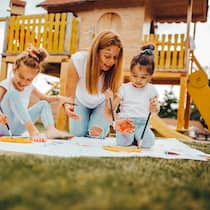 Das Abc des Spielens Zwei Kinder im Freien auf einer Wiese malen mit Farbe auf ein Blatt Papier und haben zusammen mit ihrer Mutter sichtlich Spass am Spielen.