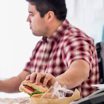 Ohne Stress abnehmen Ein übergewichtiger Mann ist nebenbei ein Sandwich, während er am Computer arbeitet.
