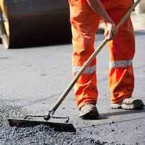 In Bern gibts eins auf die Nuss Ein Strassenbauarbeiter verteilt frischen Asphalt auf einer Strasse – Visionär und vorbildlich: Bern reguliert Lärm auf der Strasse durch Cashew-Nussschalen.