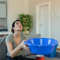 Jetzt aber hopp, Herr Vermieter! Eine Mieterin sitzt in der Wohnung auf ihrem Sofa und hält einen blauen Plastikeimer in den Händen, um Wasser von einem tropfenden Leck aufzufangen.