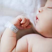Wie ändert man den Familiennamen? Ein Neugeborenes liegt auf der Geburtsstation. Das Baby schläft tief und trägt am rechten Handgelenk einen Zettel mit Geburtsdaten.