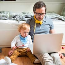 Wie steuern Chefs gut fern? Ein Vater, der im Berufsleben normalerweise Mitarbeiter führt, sitzt zu Hause auf dem Fussboden, neben ihm sein Sohn, der ebenfalls auf den Bildschirm des Laptops schaut.