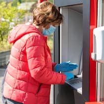 Ist mein erspartes Geld sicher? Frau mit Maske steht vor einem Geldautomaten und bezieht Geld.