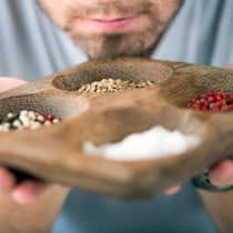 Heute schon am Pfeffer geschnuppert? Vier verschiedene Gewürze sind auf einem Holzteller verteilt. Ein Mann riecht an den Gewürzen.