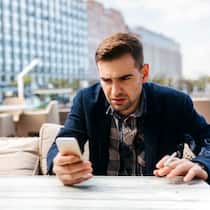 Weniger Ärger mit dem Telefon Ein Mann sitzt auf einer Terrasse im Restaurant und schaut leicht genervt auf sein Handy während er eine Zigarette raucht.