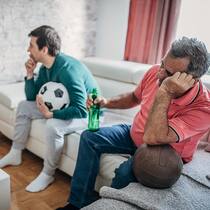Wie sollen Kinder damit umgehen? Ein Vater und sein Sohn schauen sich zusammen ein Fussballspiel an, während auf dem Wohnzimmertisch mehrere Flaschen mit Alkohol herumstehen. Dabei sieht der Vater sichtbar benebelt aus.