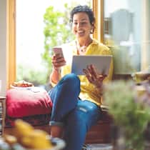 Aus der Ferne auf mobile Geräte zugreifen Eine Frau sitzt entspannt zu Hause auf dem Fenstersims und hält in ihrer linken Hand ein Tablet, in der rechten ein Smartphone.