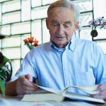 Wenn der Vater sich mehr und mehr zurückzieht Alter Mann in blauem Hemd liest vor einem Fenster ein Buch an einem Tisch mit Blumen. Stirbt ein Elternteil, und sind die Kinder bereits erwachsen und aus dem Haushalt ausgezogen, kann sich die hinterbliebene Person schnell einsam fühlen. Wie können Angehörige helfen?