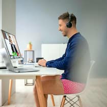 10 einfache Tipps für Videokonferenzen und Chats Ein junger Geschäftsmann im Freizeitlook sitzt zu Hause vor dem Computer und unterhält sich per Videochat mit einem Arbeitskollegen.