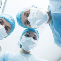 Neuer Job – soll ich die Operation verschieben? Zwei Chirurgen und eine Chirurgin stehen in einem Operationssaal und blicken von oben auf den vermeintlichen Patienten herab.