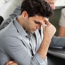 Darf mein Chef die Krankheit erwähnen? Ein Mann stützt seinen Kopf an seine Hand vor einem Tisch mit arbeitenden Personen. Darf der Chef eine Krankheit im Arbeitszeugnis erwähnen?