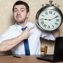 Bestellt, bezahlt – und nicht geliefert Mann sitzt an einem Schreibtisch und zeigt auf eine Wanduhr, die er in der Hand hält.