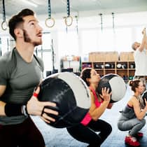 So werden Sie die Extrapfunde wieder los Eine Gruppe junger Menschen steht in einem Fitnessraum und macht Kraftübungen mit schweren Bällen. Um Gewicht zu verlieren, gibt es einige Tipps in Sachen Ernährung. Doch Abnehmen funktioniert nicht ohne sportliche Betätigung.