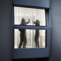 Wann schreitet die Polizei ein? Durch ein Fenster mit Vorhängen sieht man die verschwommenen Konturen eines sich streitenden Paares. Der Mann erhebt seine Hand und will offensichtlich zum Schlag ausholen, während sich die Frau mit den Armen zu schützen versucht.