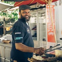 Wie Geflüchtete im Arbeitsmarkt Fuss fassen Grillmeister Baba steht am Grill beim Restaurant Pumpstation in Zürich, hält die Grillzange in der Hand und lächelt in die Kamera.