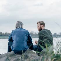 Soll ich mich mit meinem Vater treffen? Junger Mann sitzt mit seinem Vater am Ufer eines Sees und schaut den Vater an