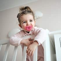 Soll man Kinderwünsche erfüllen? Ein kleines Mädchen steht in ihrem Kinderbett mit Nuggi im Mund und schaut sehnsüchtig nach ihren Eltern.