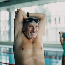 So schafft man es, dass sich die Muskeln nicht zurückbilden Ein älterer Mann und eine ältere Frau stehen am Beckenrand einer Schwimmhalle und wärmen sich mit Dehnübungen auf.